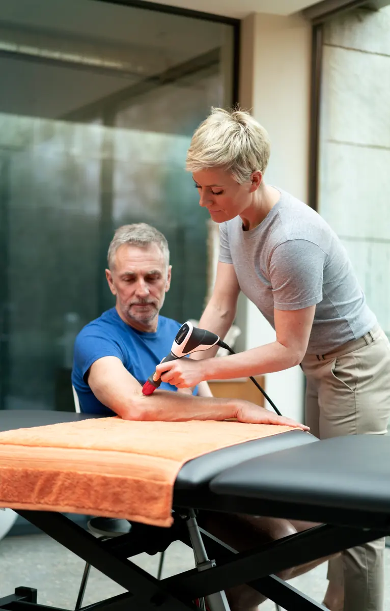 Clinician demonstrating a radial pressure wave handpiece around a seated patient’s arm